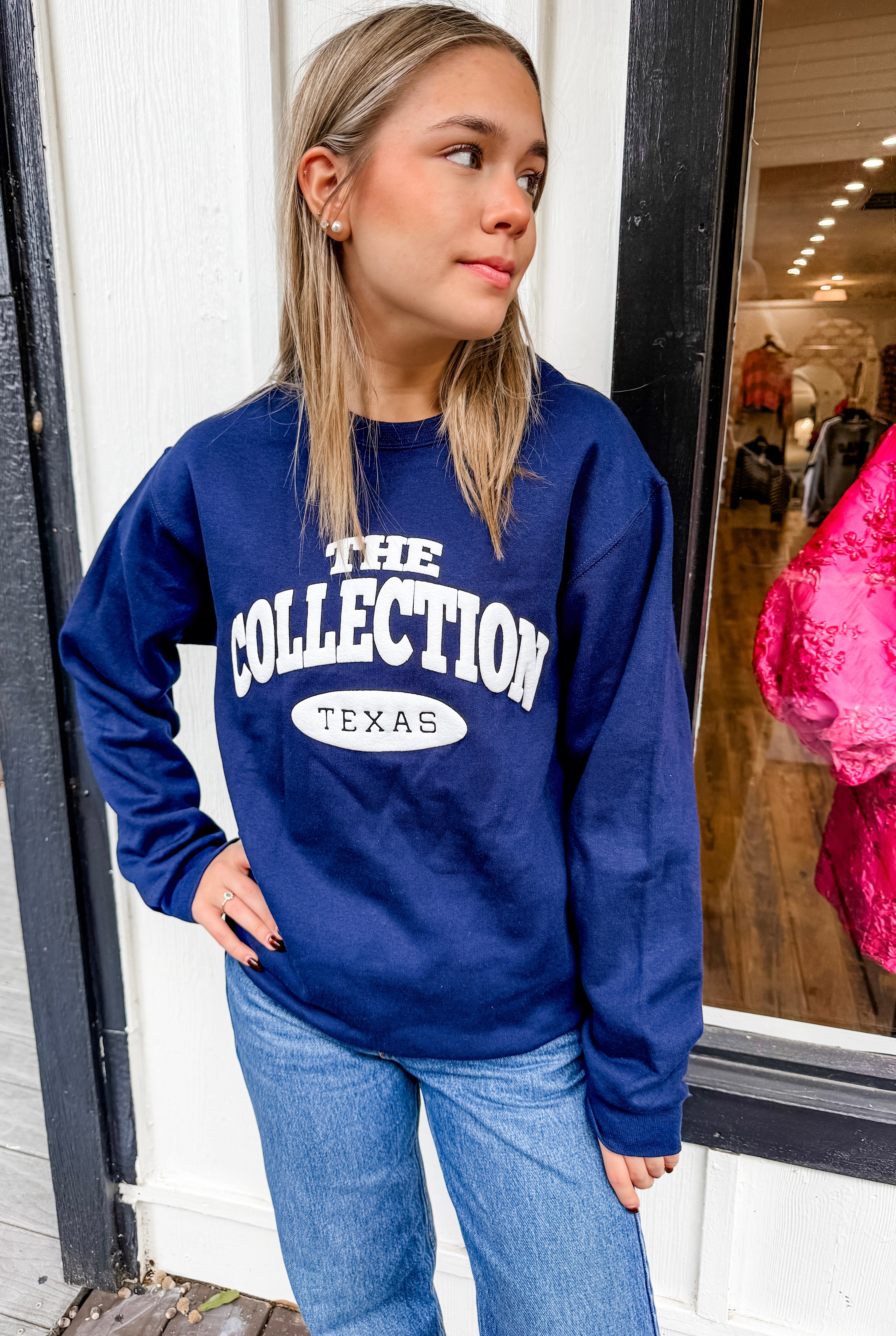 Person wearing a blue sweatshirt with 'The Collection Texas' text, standing in front of a store window.